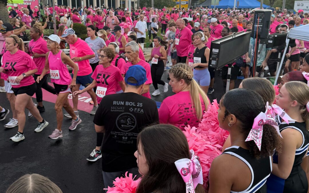 A large group of people in pink shirts participate in a race, crossing a "START/FINISH" line Cheerleaders with pink bows and pom poms stand in the foreground, and palm trees are visible in the background