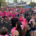 A large group of people in pink shirts participate in a race, crossing a "START/FINISH" line Cheerleaders with pink bows and pom poms stand in the foreground, and palm trees are visible in the background