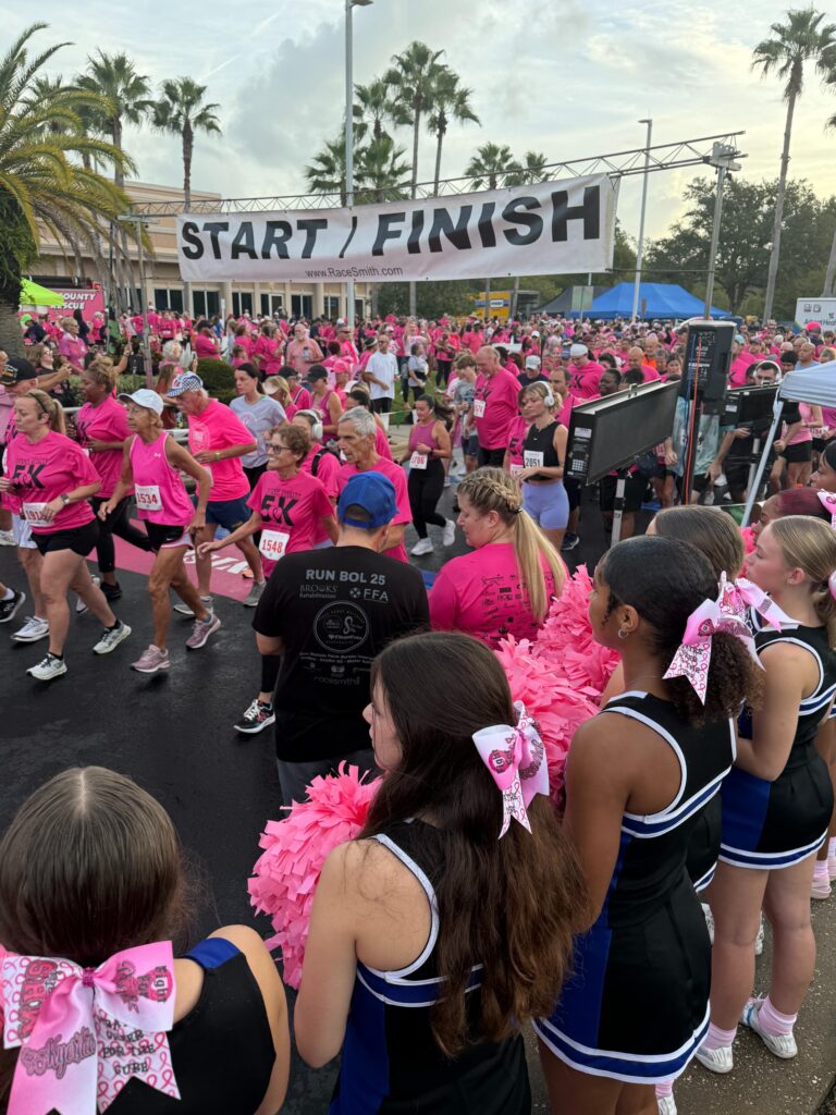 A large group of people in pink shirts participate in a race, crossing a "START/FINISH" line Cheerleaders with pink bows and pom poms stand in the foreground, and palm trees are visible in the background