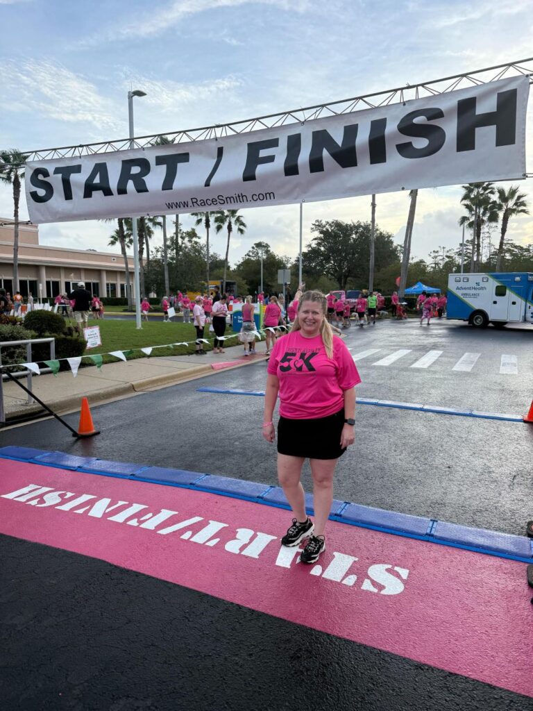 A woman in a bright pink K shirt stands and smiles at the start/finish line of a race, with other participants in matching shirts, an ambulance, and palm trees visible in the background