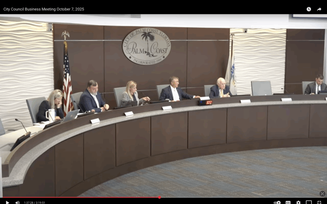 Five people sit at a curved desk during a Palm Coast City Council meeting The city’s seal is displayed on the wall behind them American and city flags stand to the side Each person has a microphone and paperwork