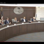 Five people sit at a curved desk during a Palm Coast City Council meeting The city’s seal is displayed on the wall behind them American and city flags stand to the side Each person has a microphone and paperwork