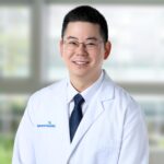 A male doctor with glasses and short black hair, wearing a white lab coat with the AdventHealth logo and a dark tie, stands smiling in a bright medical office with large windows in the background