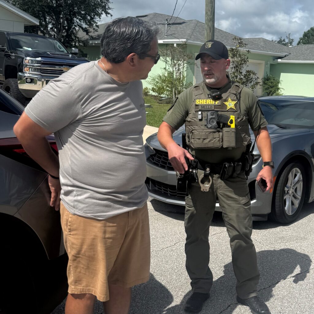 A man in a gray shirt and tan shorts talks to a sheriff’s deputy wearing a uniform, tactical vest, and cap on a residential street next to parked cars Houses and trees are visible in the background on a sunny day A man in a gray shirt and tan shorts talks to a sheriff’s deputy wearing a uniform, tactical vest, and cap on a residential street next to parked cars Houses and trees are visible in the background on a sunny day