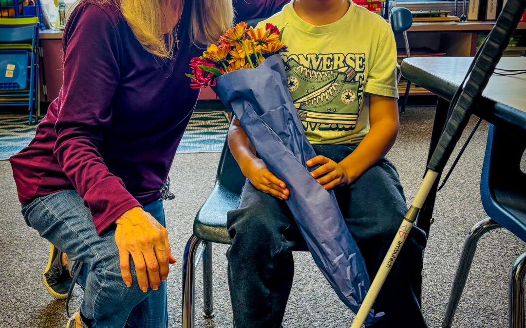 A smiling woman kneels beside a young boy sitting on a chair, holding a bouquet of flowers wrapped in blue paper and a white cane They are indoors, with shelves and windows in the background