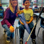 A smiling woman kneels beside a young boy sitting on a chair, holding a bouquet of flowers wrapped in blue paper and a white cane They are indoors, with shelves and windows in the background