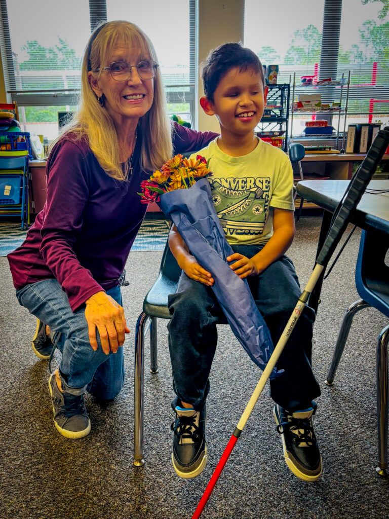 A smiling woman kneels beside a young boy sitting on a chair, holding a bouquet of flowers wrapped in blue paper and a white cane They are indoors, with shelves and windows in the background