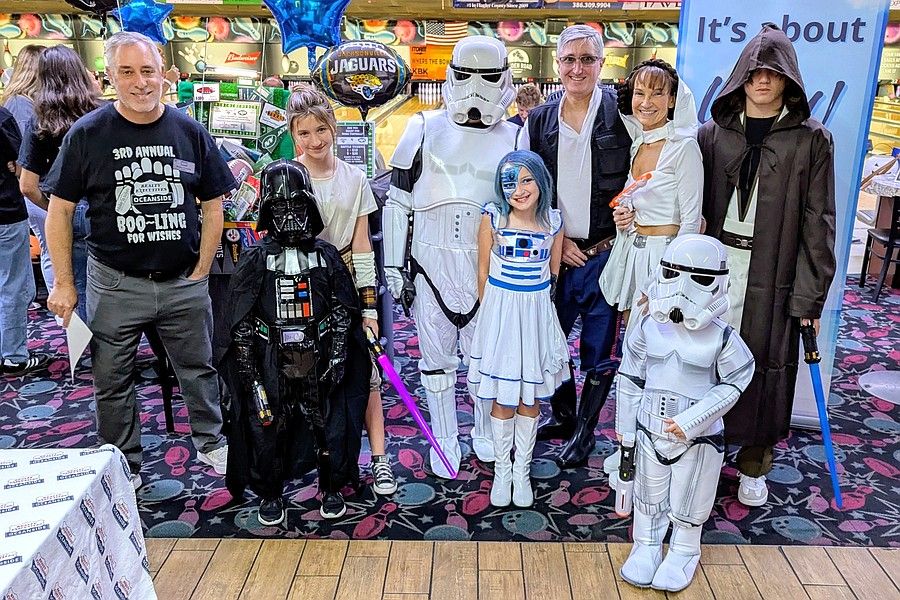 A group of people, adults and children, dressed in Star Wars costumes including Darth Vader, Stormtroopers, R D, Han Solo, Princess Leia, and a Jedi, pose together in a brightly decorated indoor space A group of people, adults and children, dressed in Star Wars costumes including Darth Vader, Stormtroopers, R D, Han Solo, Princess Leia, and a Jedi, pose together in a brightly decorated indoor space