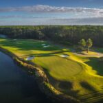 Aerial view of a golf course with bright green fairways and putting greens beside a large body of water, bordered by dense trees under a partly cloudy sky