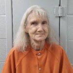 An older woman with long, light gray hair and a slight smile wears an orange prison jumpsuit, seated against a light colored cinderblock wall and a metal door