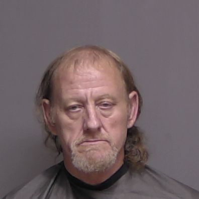 A middle aged man with light skin, thinning light brown hair, and a goatee, wearing a dark garment, poses against a plain, gray background for a mugshot style photo