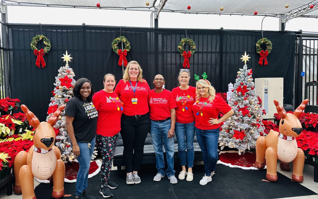 Six people, true Community Heroes, stand smiling in front of white Christmas trees, wreaths, red poinsettias, and inflatable reindeer Five wear matching red shirts and one a black shirt, creating a cheerful and festive scene