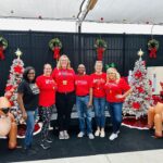 Six people, true Community Heroes, stand smiling in front of white Christmas trees, wreaths, red poinsettias, and inflatable reindeer Five wear matching red shirts and one a black shirt, creating a cheerful and festive scene