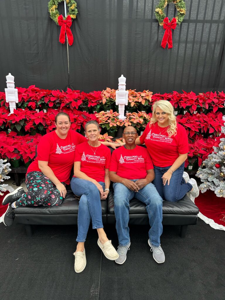 Four Community Heroes wearing matching red Christmas shirts sit and pose on a black bench, surrounded by red poinsettias, white candlestick decorations, and holiday wreaths against a festive backdrop