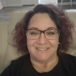 A woman with wavy, shoulder length hair and glasses smiles while sitting on a light colored chair in a modern, well lit room with visible doors and a kitchen in the background