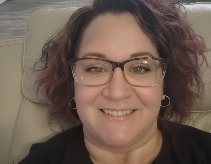 A woman with wavy, shoulder length hair and glasses smiles while sitting on a light colored chair in a modern, well lit room with visible doors and a kitchen in the background