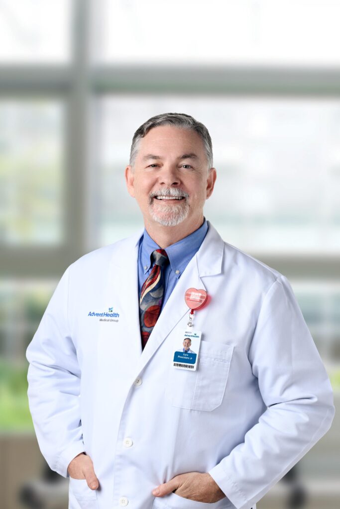 A middle aged male doctor with gray hair and a beard smiles while wearing a white lab coat, blue shirt, patterned tie, and ID badge He stands in a bright, blurred indoor setting