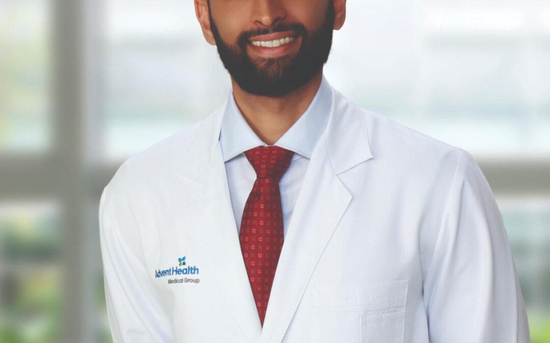 A man with a beard wearing a white lab coat, red tie, and light blue shirt stands and smiles in front of a blurred background with large windows