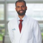 A man with a beard wearing a white lab coat, red tie, and light blue shirt stands and smiles in front of a blurred background with large windows