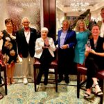 Eight adults, five standing and three seated, pose indoors with smiles The three seated women each hold glass awards The group is dressed in formal or business attire, and the setting appears to be a hotel or event venue