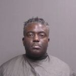 A man with short, gray and black hair wearing a dark colored smock poses for a mugshot against a plain, neutral background
