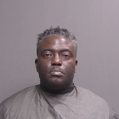 A man with short, gray and black hair wearing a dark colored smock poses for a mugshot against a plain, neutral background