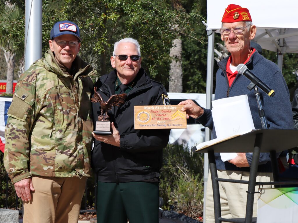 Three older men stand outside at an event One holds an eagle trophy and a plaque, another stands at a podium, and the third is dressed in camouflage They are smiling and appear to be at an awards ceremony or presentation