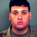 A young man with short, dark curly hair wearing a camouflage patterned shirt stares directly at the camera with a neutral expression against a plain background