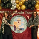 Two women in formal attire stand smiling and gesturing toward a banner that reads “Bringing People Together” and features the Hispanic American Cultural Society logo, surrounded by gold, black, and silver balloons