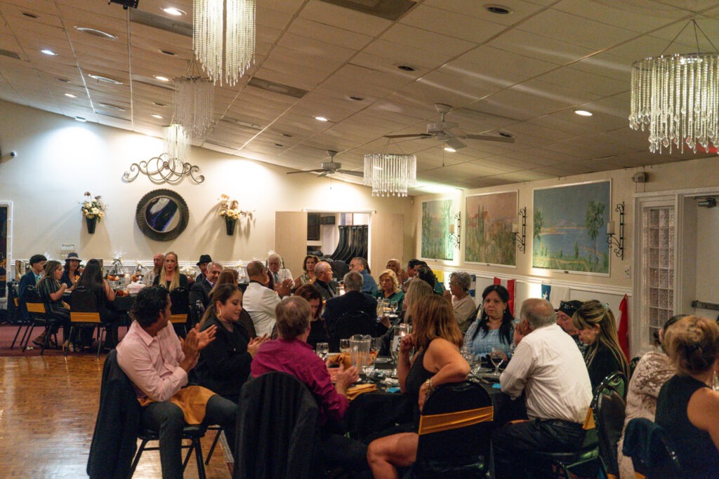 A large group of people sit around tables in a decorated banquet hall, enjoying a formal dinner event Chandeliers hang from the ceiling, and framed art decorates the walls Some guests are talking, eating, or clapping