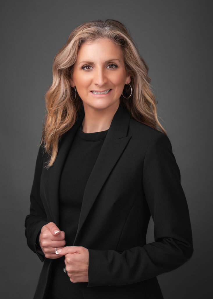 A woman with long wavy blonde hair smiles at the camera She is wearing a black blazer over a black top, with hoop earrings and a ring, standing against a dark gray background