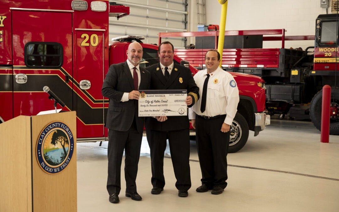 Three men in formal attire stand together, smiling and holding a large ceremonial check inside a fire station A red fire truck is visible in the background A podium with a circular seal is on the left