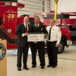 Three men in formal attire stand together, smiling and holding a large ceremonial check inside a fire station A red fire truck is visible in the background A podium with a circular seal is on the left