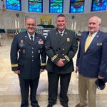 Three men stand smiling inside a church with stained glass windows One wears a military uniform, one a firefighter uniform, and one a suit with a pin An altar with candles and a U S flag is visible in the background