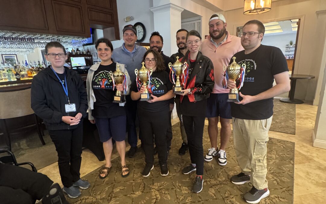 A group of nine people stand indoors, smiling and holding trophies Some are dressed casually, and some wear glasses They are posing together on a patterned carpet in a well lit room with white walls and wooden accents