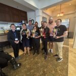 A group of nine people stand indoors, smiling and holding trophies Some are dressed casually, and some wear glasses They are posing together on a patterned carpet in a well lit room with white walls and wooden accents