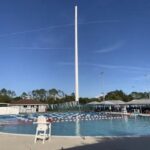 An outdoor swimming pool with lane dividers, a lifeguard chair, and a tall empty flagpole in the center Trees and buildings are visible in the background under a clear blue sky