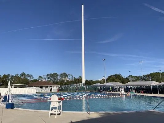 An outdoor swimming pool with lane dividers, a lifeguard chair, and a tall empty flagpole in the center Trees and buildings are visible in the background under a clear blue sky