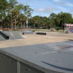 An outdoor skatepark featuring ramps, rails, and platforms, surrounded by trees and a fence The concrete surface is clear, and some ramps have graffiti The sky is mostly clear with scattered clouds