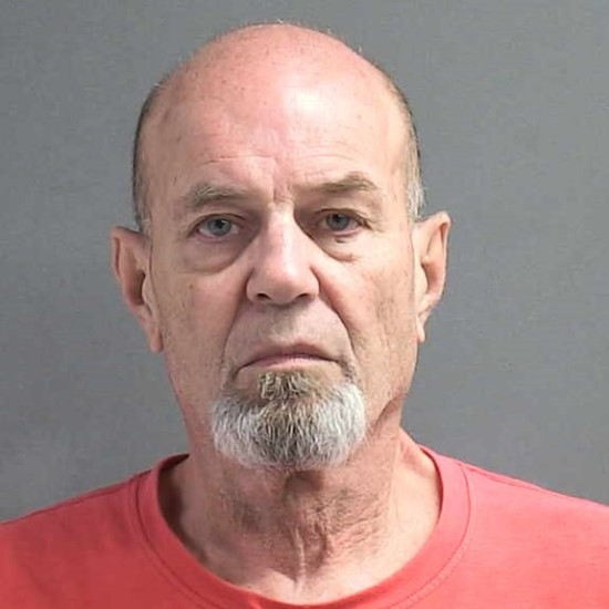 A bald older man with a gray goatee and mustache is wearing a red shirt, looking straight at the camera with a neutral expression, against a plain gray background