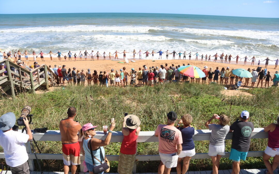 A large group of people form a human chain along a sandy beach near the shoreline, while others watch and take photos from a wooden boardwalk shaded by umbrellas, with waves in the background under a clear sky