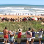 A large group of people form a human chain along a sandy beach near the shoreline, while others watch and take photos from a wooden boardwalk shaded by umbrellas, with waves in the background under a clear sky