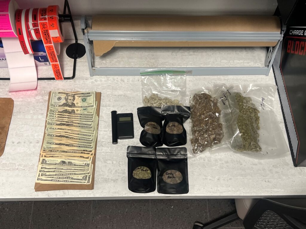 A table with stacks of cash, a small digital scale, plastic bags of marijuana, black pouches labeled with strains, and rolls of shipping labels