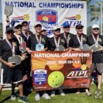 A group of eight men wearing medals and holding pickleball paddles stand together, smiling, behind a banner reading “NATIONAL CHAMPIONS : MEN + ” A backdrop for the American Team Pickleball League is visible behind them