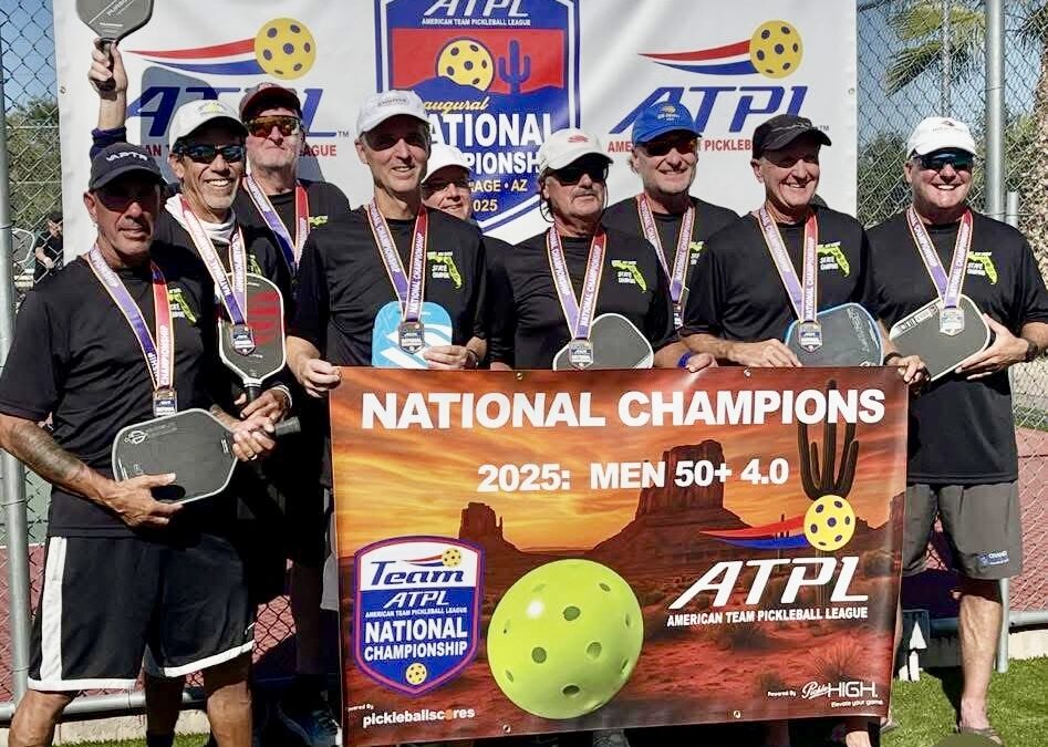 A group of eight men wearing medals and holding pickleball paddles stand together, smiling, behind a banner reading “NATIONAL CHAMPIONS : MEN + ” A backdrop for the American Team Pickleball League is visible behind them