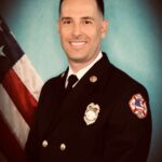 A firefighter in formal dress uniform stands smiling in front of an American flag, wearing a badge, decorative buttons, and a firefighter patch on the sleeve, with a blue background