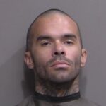 A man with a shaved head and facial tattoos looks directly at the camera with a neutral expression against a plain background