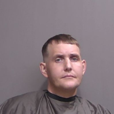 A man with short brown hair wearing a dark colored garment poses against a plain gray background