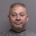 A middle aged man with short, thinning hair and facial stubble is facing the camera against a plain background, wearing a dark, protective garment