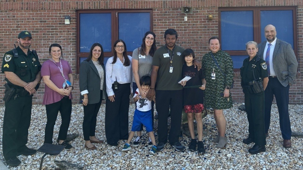 A group of eleven people, including two uniformed officers, several adults in business or casual attire, and two children, stand smiling together outside in front of a brick building with blue windows